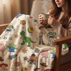 May include: A cream-colored blanket featuring cartoon characters from a popular animated film. The blanket has images of a green dinosaur, a space ranger, a cowboy, and other characters. The blanket is decorated with stars and other shapes.