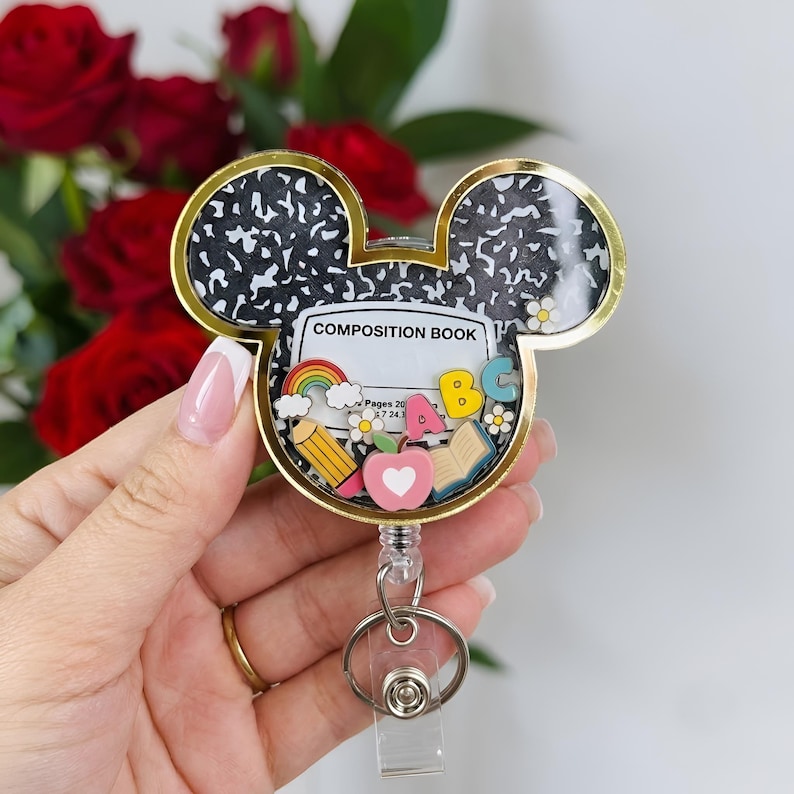 May include: A Mickey Mouse-shaped badge reel featuring a black and white speckled composition book pattern. The center displays a white label with the text COMPOSITION BOOK, surrounded by small colorful charms including a rainbow, pencil, apple, book, and letters.