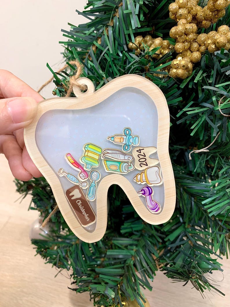 May include: Wooden tooth-shaped ornament with a clear center filled with miniature dental tools and the year 2024. The ornament has a light brown wooden frame and a small tag that says "Christopher".