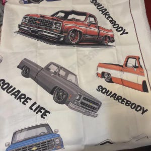 May include: White fabric featuring illustrations of classic pickup trucks in various colors, including red, gray, and orange. The words "SQUAREBODY" and "SQUARE LIFE" are printed on the fabric, which is likely intended for crafting or apparel.