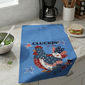May include: A blue waffle-weave kitchen towel with a patriotic chicken design. The chicken is made up of red, white, and blue patterns, including stars and stripes. The text "CLUCKIN' in the U.S." is printed above the chicken.