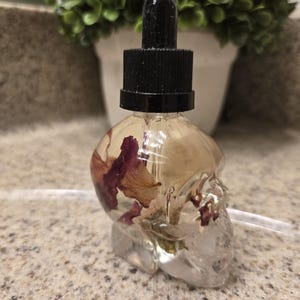 Könnte beinhalten: Eine durchsichtige Glasflasche in Schädelform, gefüllt mit einer hellgelben Flüssigkeit und getrockneten rosa Blüten. Die Flasche hat einen schwarzen Tropferaufsatz.