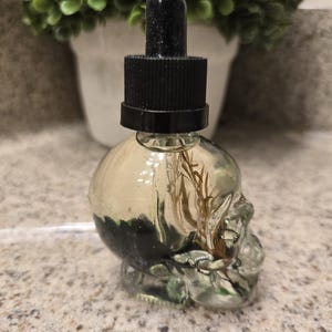 Puede incluir: Una botella de vidrio transparente con forma de cráneo con un tapón cuentagotas negro. La botella está llena de un líquido transparente y contiene material vegetal seco.