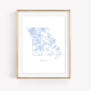 Puede incluir: Una impresión enmarcada que presenta el contorno del estado de Missouri, lleno de un delicado patrón floral azul. La palabra "Missouri" está escrita en cursiva debajo. El marco es dorado.