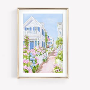 以下が含まれることがあります： 白い家、青い窓、鮮やかなピンクと青のアジサイが並ぶ、趣のある通りの水彩画の額入りプリント。白い柵が歩道を縁取っています。アメリカの国旗が家の1つからぶら下がっています。