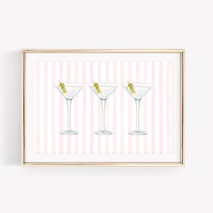 May include: A framed print featuring three martini glasses, each garnished with an olive. The glasses are set against a pink and white striped background. The frame is gold.