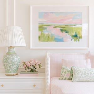 Op de afbeelding: Een ingelijst landschapsschilderij met een moeraslandschap met roze en blauwe luchtreflecties in het water, omringd door groen gras. Het kunstwerk hangt boven een wit nachtkastje met een groene bloemenlamp en een boeket roze rozen.