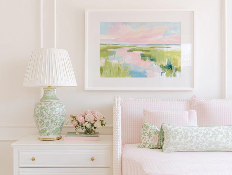 Op de afbeelding: Een ingelijst landschapsschilderij met een moeraslandschap met roze en blauwe luchtreflecties in het water, omringd door groen gras. Het kunstwerk hangt boven een wit nachtkastje met een groene bloemenlamp en een boeket roze rozen.