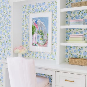 May include: A home office featuring a white desk, chair, and shelving unit. The walls are adorned with blue floral wallpaper. A framed watercolour painting of a house with an American flag hangs above the desk. A vase holds pink and white flowers.