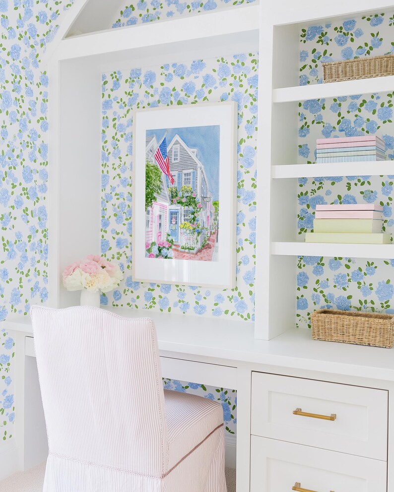 May include: A home office featuring a white desk, chair, and shelving unit. The walls are adorned with blue floral wallpaper. A framed watercolour painting of a house with an American flag hangs above the desk. A vase holds pink and white flowers.
