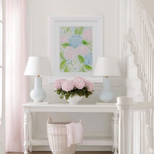 May include: A white console table featuring two light blue lamps with white shades, a central white bowl filled with pink hydrangea flowers, and a white wicker basket underneath. A framed floral painting hangs above the table on a white wall.