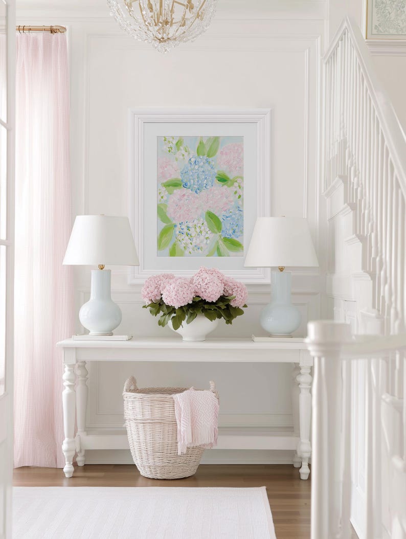 May include: A white console table featuring two light blue lamps with white shades, a central white bowl filled with pink hydrangea flowers, and a white wicker basket underneath. A framed floral painting hangs above the table on a white wall.