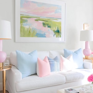 Op de afbeelding: Een witte bank met vijf decoratieve kussens in lichtblauw, effen roze en een blauw-wit ruitpatroon. Boven de bank hangt een ingelijst landschapsschilderij met roze, groene en blauwe penseelstreken dat een moeraslandschap weergeeft.