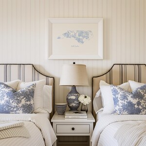 May include: Two twin beds with white and blue striped headboards and white and blue patterned pillows. A white bedside table with a lamp and a book sits between the beds. A framed print of the state of North Carolina hangs on the wall above the bedside table.