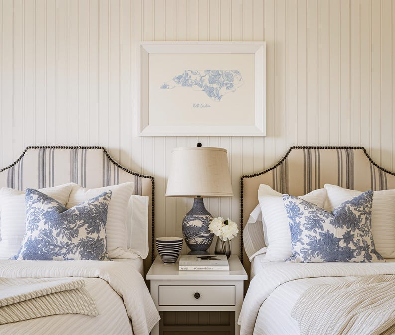 May include: Two twin beds with white and blue striped headboards and white and blue patterned pillows. A white bedside table with a lamp and a book sits between the beds. A framed print of the state of North Carolina hangs on the wall above the bedside table.