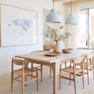 May include: A light blue floral print of the state of North Carolina hangs on a white wall above a wooden dining table with six chairs. Two light blue pendant lights hang above the table.
