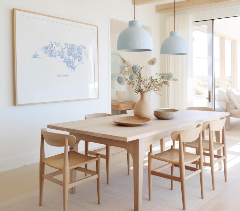 May include: A light blue floral print of the state of North Carolina hangs on a white wall above a wooden dining table with six chairs. Two light blue pendant lights hang above the table.