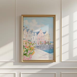 Puede incluir: Una pintura de acuarela enmarcada de un pueblo costero con edificios coloridos. La obra presenta una hilera de casas en tonos blanco, rosa y amarillo, con un cielo azul y agua. Flores en rosa, amarillo y naranja en primer plano.