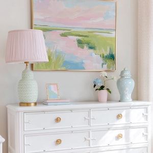Op de afbeelding: Een witte ladekast met zes lades en gouden bloemvormige knoppen, met daarop een roze geplooide lamp, een kleine fotolijst, een roze vaas met blauwe en witte hortensia's en een lichtblauwe pot met deksel, onder een ingelijst landschapsschilderij.