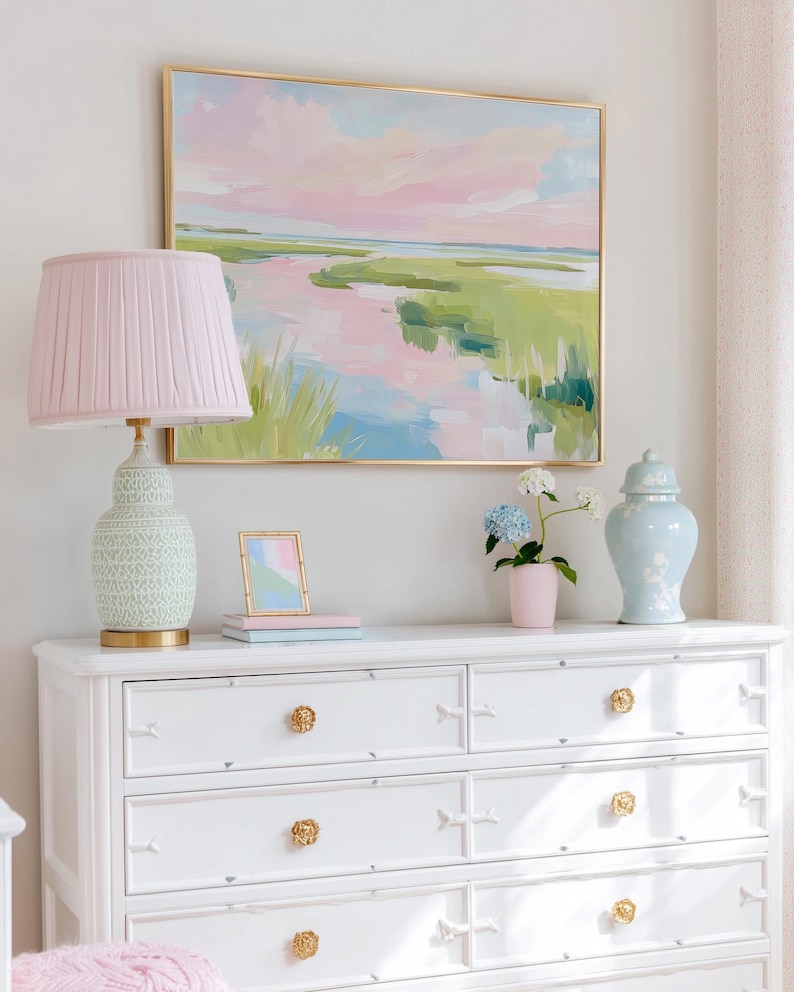 Op de afbeelding: Een witte ladekast met zes lades en gouden bloemvormige knoppen, met daarop een roze geplooide lamp, een kleine fotolijst, een roze vaas met blauwe en witte hortensia's en een lichtblauwe pot met deksel, onder een ingelijst landschapsschilderij.