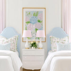 May include: Two light blue upholstered headboards with white piping, white bedding, and patterned pillows. A white three-drawer nightstand sits between them, holding two white lamps with pink shades and a small vase of white flowers under a floral art print.