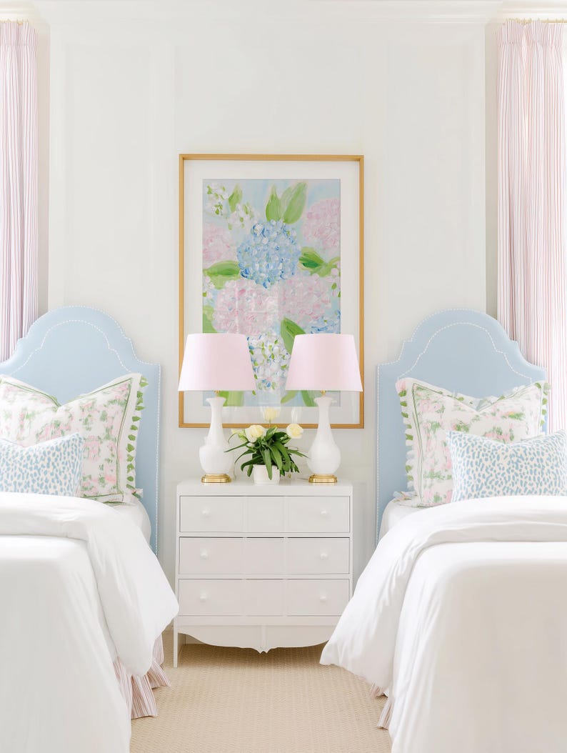 May include: Two light blue upholstered headboards with white piping, white bedding, and patterned pillows. A white three-drawer nightstand sits between them, holding two white lamps with pink shades and a small vase of white flowers under a floral art print.