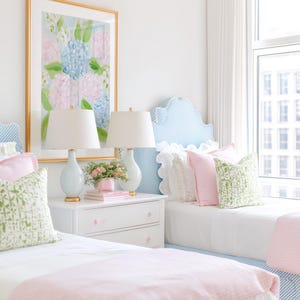 May include: A bedroom featuring two twin beds with blue and white striped headboards, white bedding, and pink blankets. A white nightstand sits between the beds, holding two light blue lamps and a small pink vase with flowers under a floral art print.
