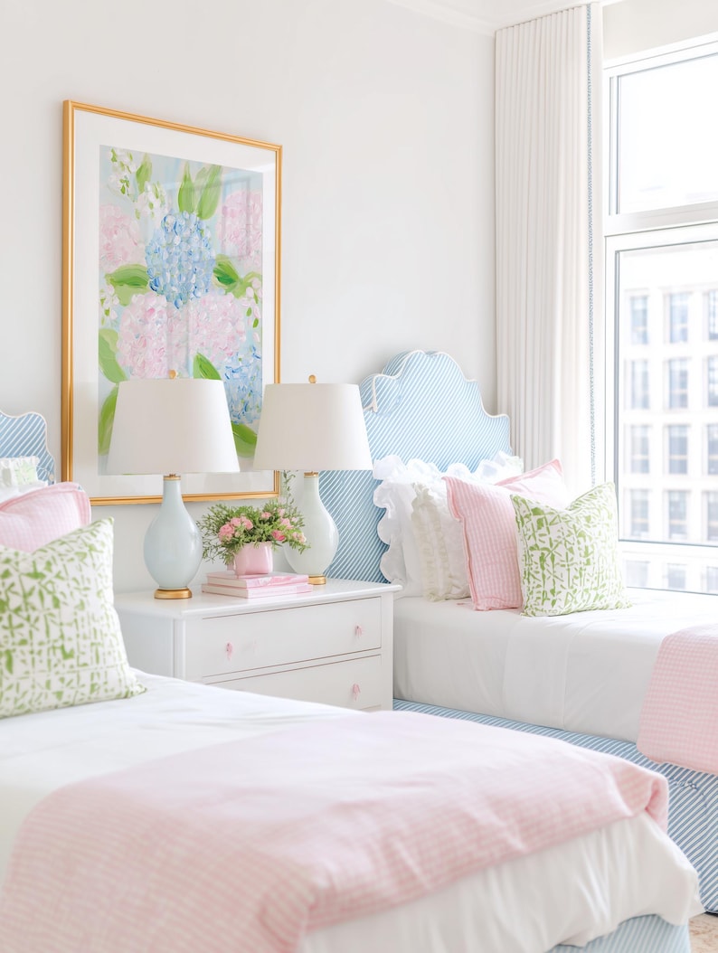 May include: A bedroom featuring two twin beds with blue and white striped headboards, white bedding, and pink blankets. A white nightstand sits between the beds, holding two light blue lamps and a small pink vase with flowers under a floral art print.