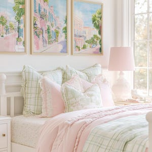 May include: A bedroom scene featuring a white bed frame with a pink comforter and green plaid blanket. Decorative pillows in pink and green hues adorn the bed. Above the bed, three framed watercolor paintings depict a street scene with pink buildings and palm trees.
