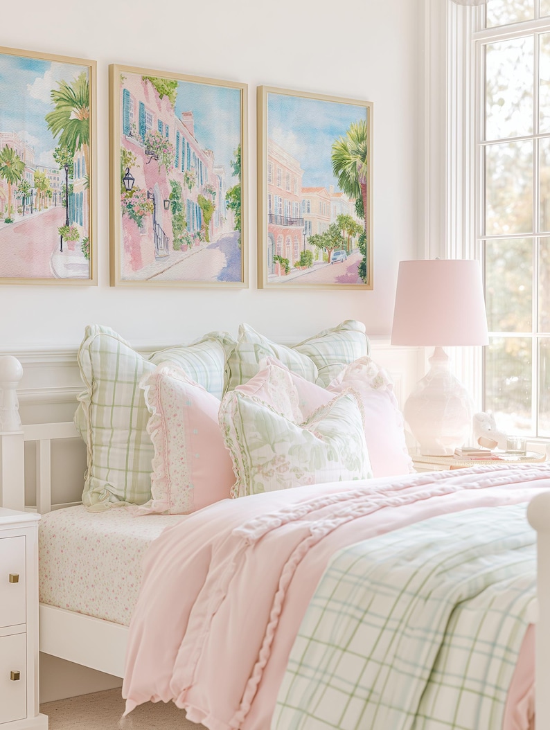 May include: A bedroom scene featuring a white bed frame with a pink comforter and green plaid blanket. Decorative pillows in pink and green hues adorn the bed. Above the bed, three framed watercolor paintings depict a street scene with pink buildings and palm trees.