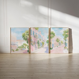 May include: Set of three framed watercolor prints depicting a charming street scene. The artwork features pink buildings with blue shutters, palm trees, and a light blue sky. The prints are displayed on a light wood floor against a white wall.