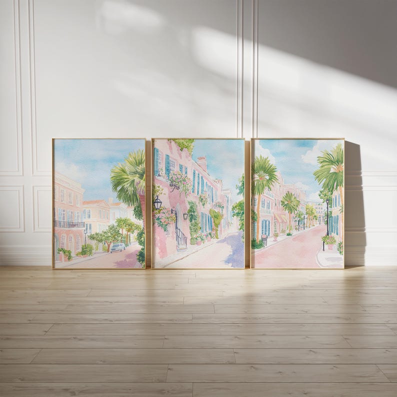 May include: Set of three framed watercolor prints depicting a charming street scene. The artwork features pink buildings with blue shutters, palm trees, and a light blue sky. The prints are displayed on a light wood floor against a white wall.