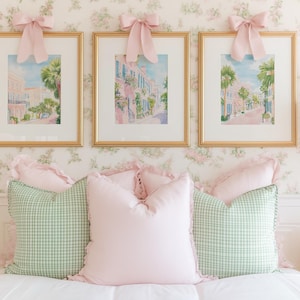 May include: Three framed watercolor prints of a street scene with pink buildings and palm trees, each topped with a pink bow. Below, a bed is adorned with pink and green gingham pillows, creating a soft, feminine aesthetic.