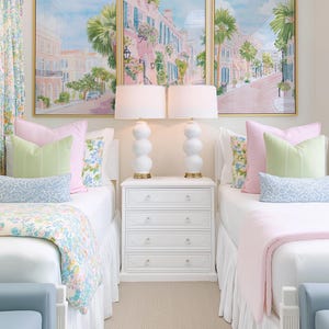 May include: A bedroom scene featuring two twin beds with white bedding, pink and green pillows, and floral patterned blankets. Above the beds are three framed paintings of a street scene. White lamps with gold bases sit on a white nightstand.