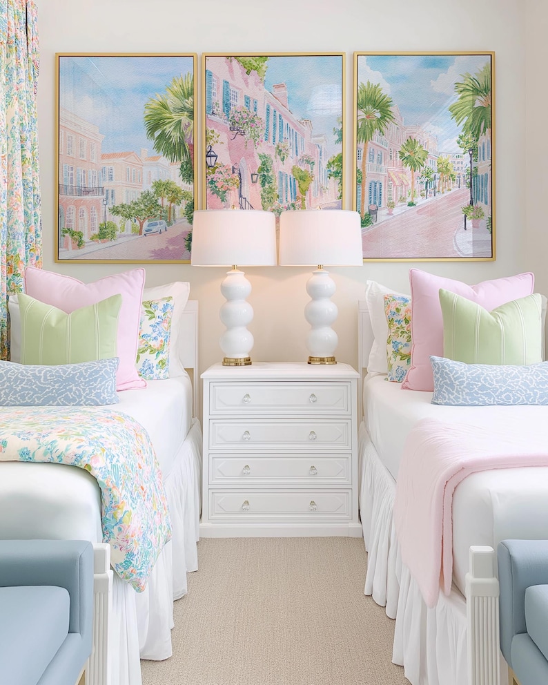 May include: A bedroom scene featuring two twin beds with white bedding, pink and green pillows, and floral patterned blankets. Above the beds are three framed paintings of a street scene. White lamps with gold bases sit on a white nightstand.