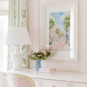 May include: A white desk with a white lamp, a blue vase with flowers, and a framed watercolor painting of a street scene. The room has pink walls, a striped curtain, and a striped chair.