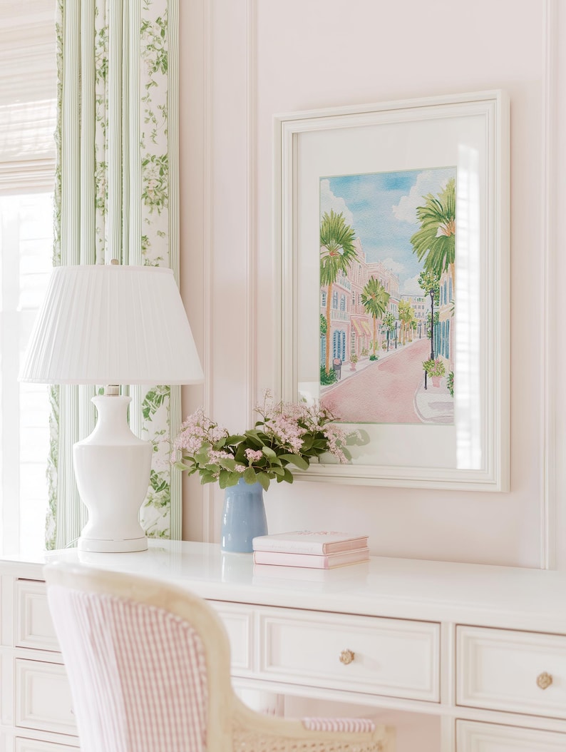 May include: A white desk with a white lamp, a blue vase with flowers, and a framed watercolor painting of a street scene. The room has pink walls, a striped curtain, and a striped chair.