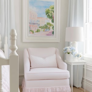 May include: A light pink striped armchair with a matching fringed skirt and a white pillow. A framed watercolor painting of a street scene hangs above the chair. A white side table holds a lamp and flowers.