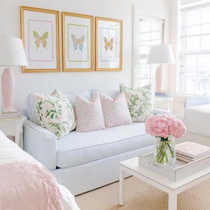 May include: A bright, airy living room scene featuring a light blue striped sofa adorned with floral and pink polka-dot pillows. Above the sofa, three framed butterfly prints in gold frames hang on the white wall. A white coffee table holds a vase of pink flowers.