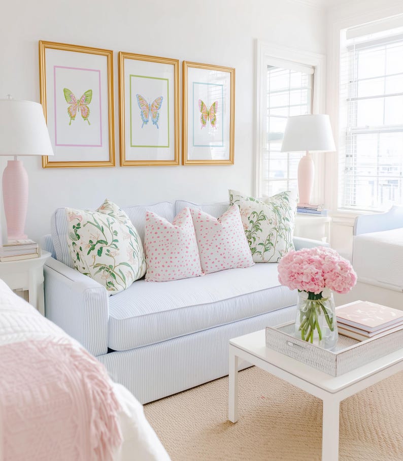 May include: A bright, airy living room scene featuring a light blue striped sofa adorned with floral and pink polka-dot pillows. Above the sofa, three framed butterfly prints in gold frames hang on the white wall. A white coffee table holds a vase of pink flowers.