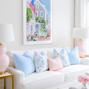 May include: A living room with a white sofa and blue and pink cushions. A framed watercolour painting of a street scene with an American flag hangs above. Pink lamps and a gold side table add to the room's decor.
