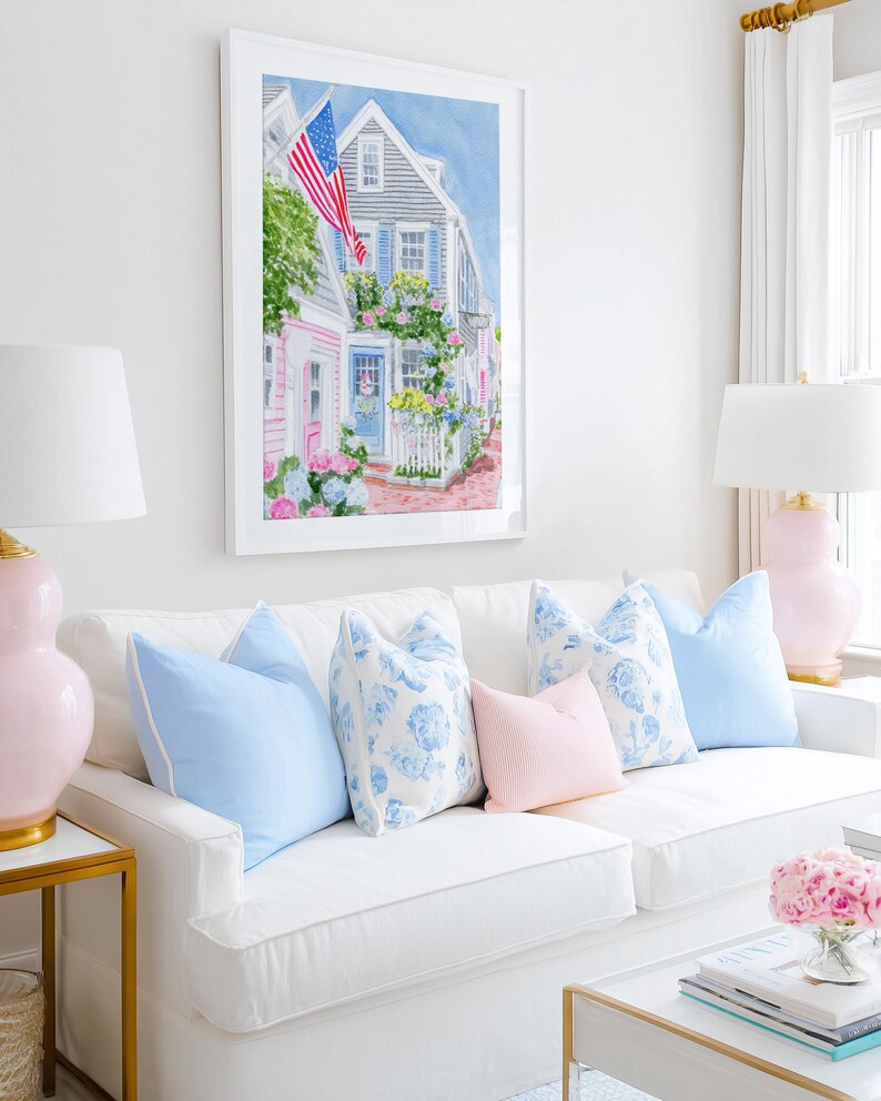 May include: A living room with a white sofa and blue and pink cushions. A framed watercolour painting of a street scene with an American flag hangs above. Pink lamps and a gold side table add to the room's decor.