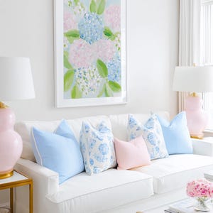 May include: A white sofa features five decorative throw pillows in solid light blue, floral blue and white, and solid pale pink. Above the sofa hangs a framed painting of blue and pink hydrangea flowers with green leaves on a light blue background.