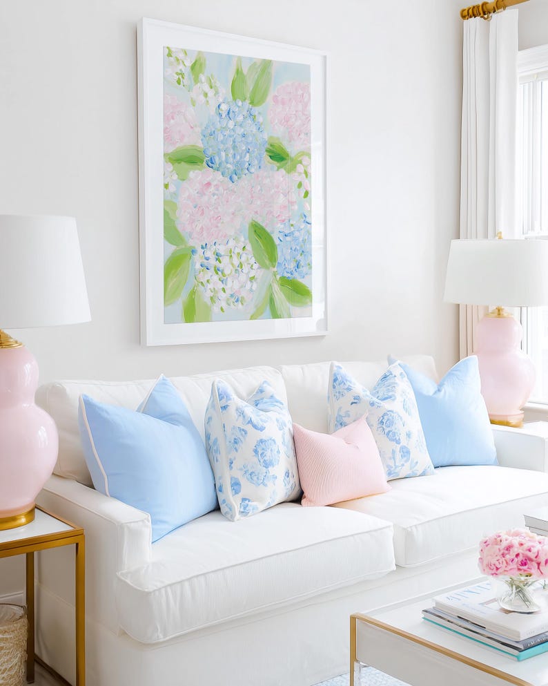 May include: A white sofa features five decorative throw pillows in solid light blue, floral blue and white, and solid pale pink. Above the sofa hangs a framed painting of blue and pink hydrangea flowers with green leaves on a light blue background.