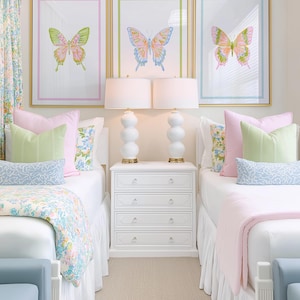 May include: Three framed butterfly art prints in gold frames, each with a different color border. The room features two beds with white bedding, pink and green pillows, and a white nightstand with lamps.