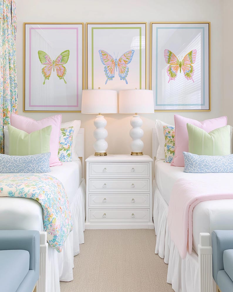May include: Three framed butterfly art prints in gold frames, each with a different color border. The room features two beds with white bedding, pink and green pillows, and a white nightstand with lamps.