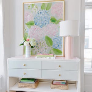 May include: A white console table with four drawers featuring gold knobs and an open bottom shelf. On top sits a framed floral painting, a striped vase with flowers, two stacked books, and a cylindrical pink lamp with a white drum shade.