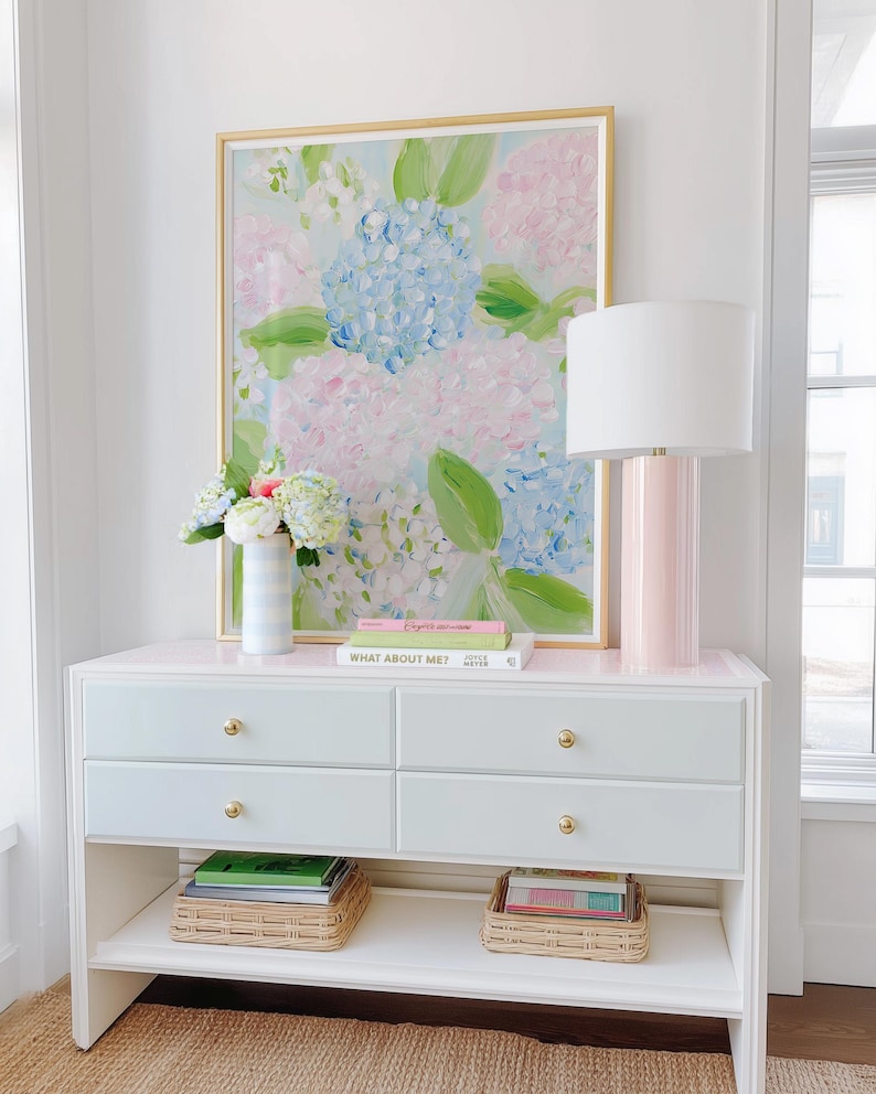 May include: A white console table with four drawers featuring gold knobs and an open bottom shelf. On top sits a framed floral painting, a striped vase with flowers, two stacked books, and a cylindrical pink lamp with a white drum shade.