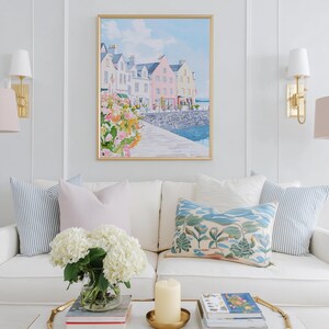 Puede incluir: Un sal&oacute;n con un sof&aacute; blanco adornado con cojines en tonos azules y rosas. Sobre el sof&aacute;, una pintura de acuarela enmarcada de un pueblo costero. L&aacute;mparas doradas y una mesa de centro con una vela y libros completan la decoraci&oacute;n.
