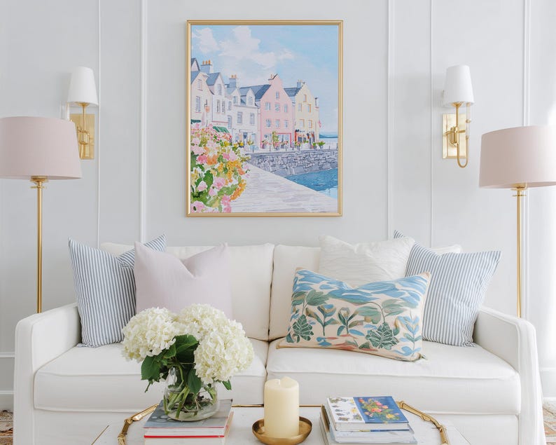 Puede incluir: Un sal&oacute;n con un sof&aacute; blanco adornado con cojines en tonos azules y rosas. Sobre el sof&aacute;, una pintura de acuarela enmarcada de un pueblo costero. L&aacute;mparas doradas y una mesa de centro con una vela y libros completan la decoraci&oacute;n.